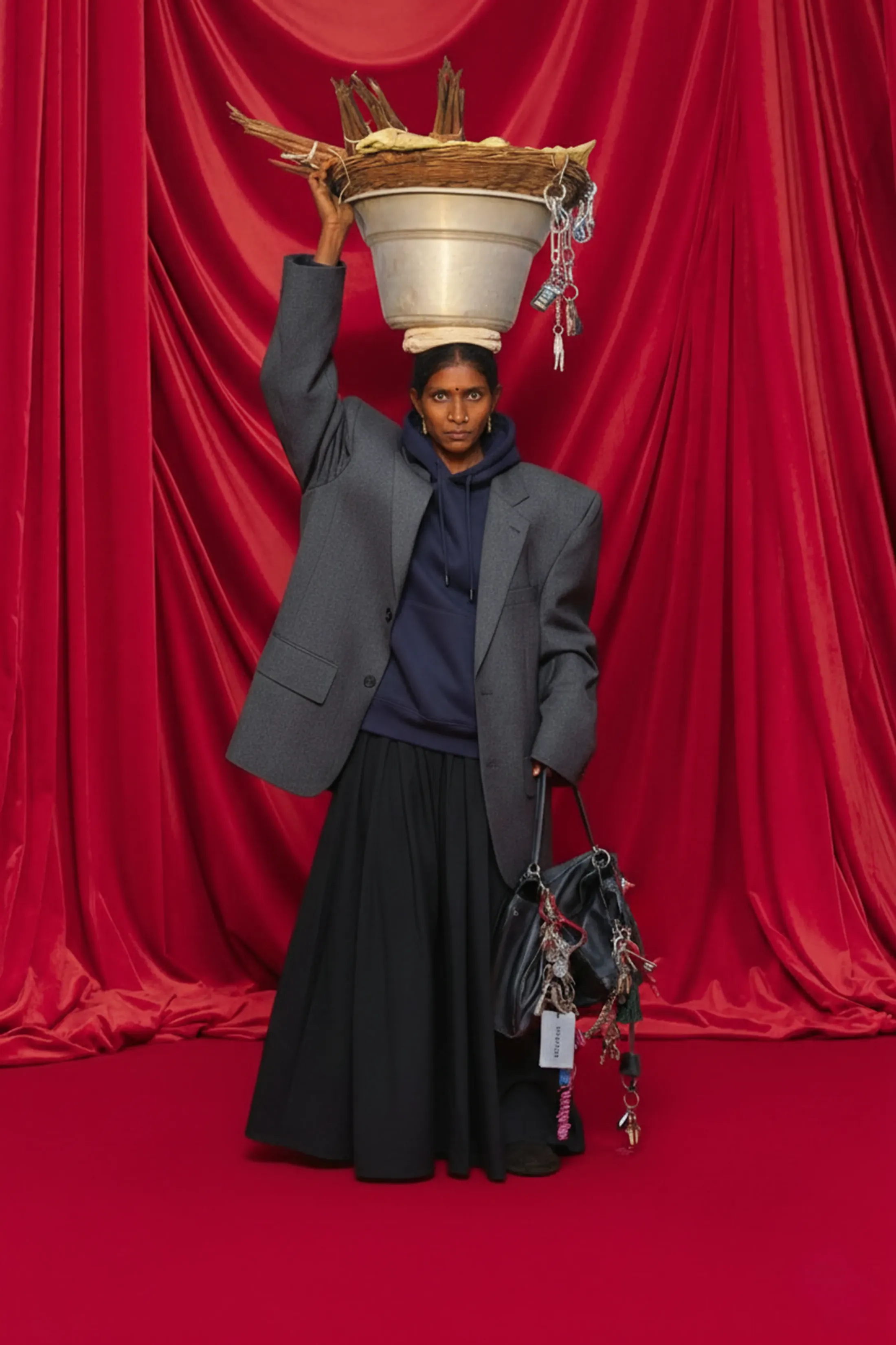 A woman in an oversized grey blazer and long black skirt against red velvet curtains, carrying a steel pot of dried grass on her head and a bag hung with charms.