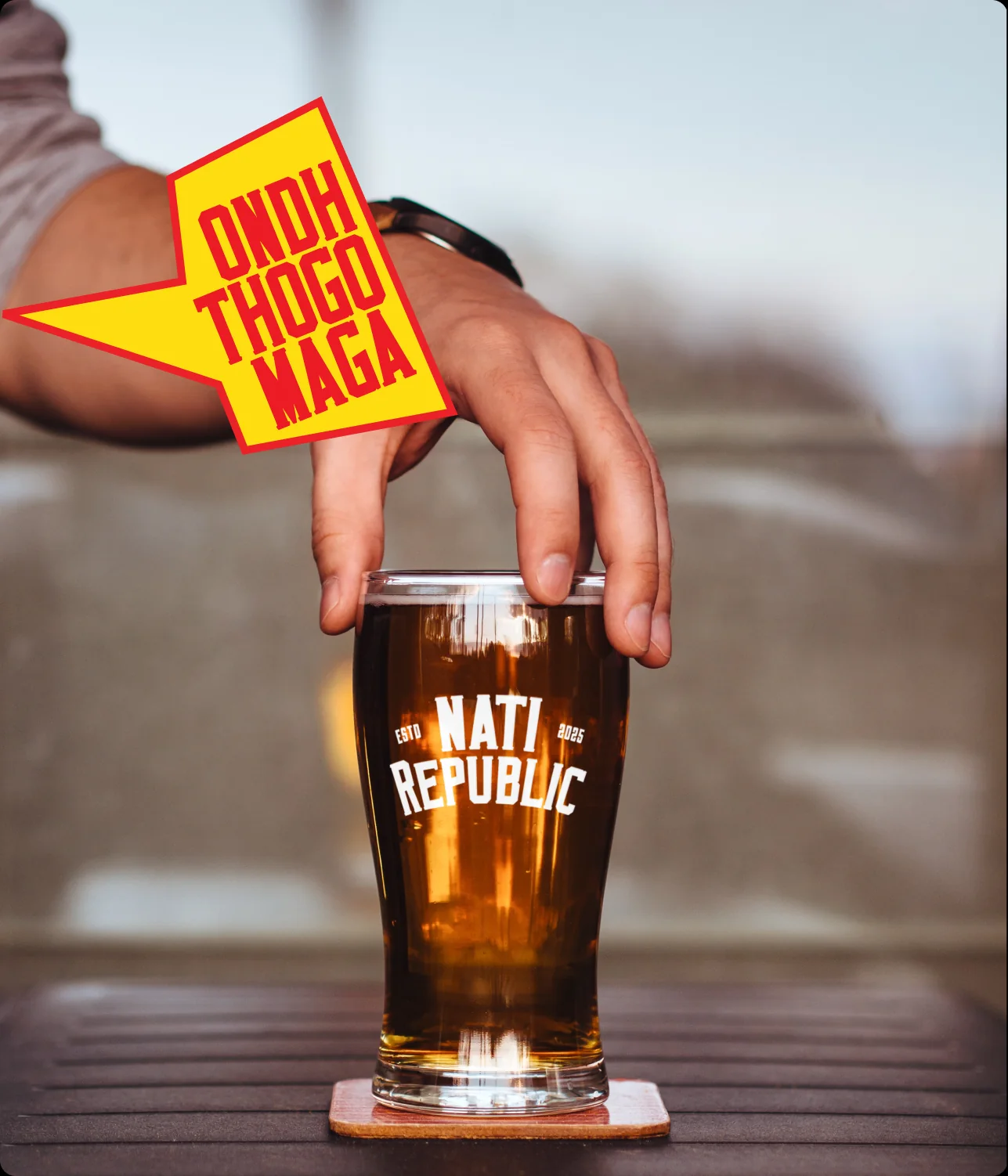 A hand setting down a Nati Republic beer glass, with a speech bubble: Ondh thogo maga.