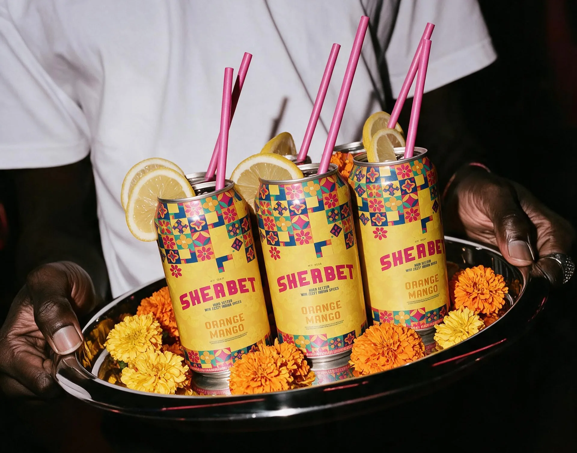 A waiter holding a steel tray of three Sherbet cans with pink straws and lemon slices, on a bed of marigolds.
